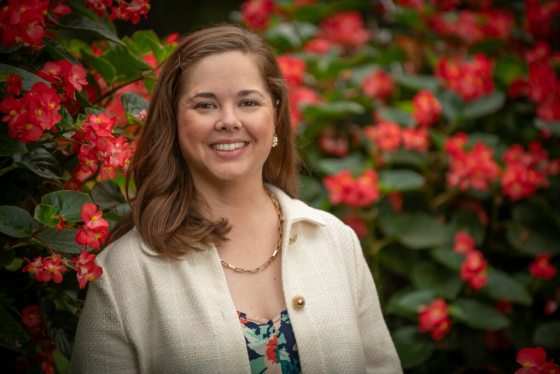 A person in a white jacket smiling in front of red flowering bushes.